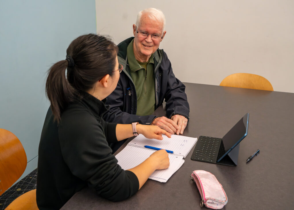 Bob and Junyi take notes on paper in their adult education session while watching a video