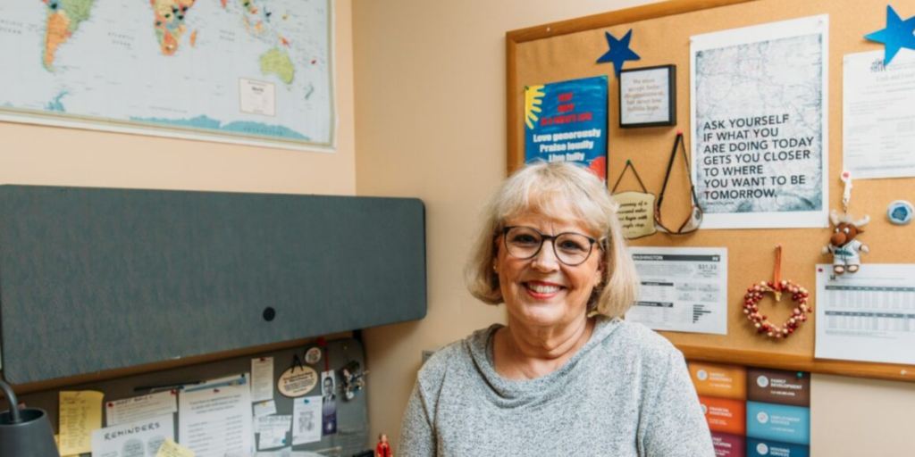 Hopelink Case Manager Linda Tappan stands in her office, smiling while looking into the camera.. Her map pin-pointing where her clients have hailed from over the years hangs above her.