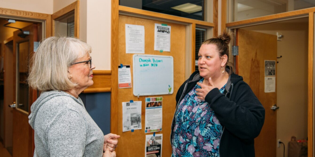 Linda chats with Family Development Program Manager Elizabeth Miller in Hopelink's Avondale Park offices. An image shows Hopelink Case Manager Linda Tappan standing to left and speaking with Family Development Program Manager Elizabeth Miller to the right in the offices of Hopelink's Avondale Park.