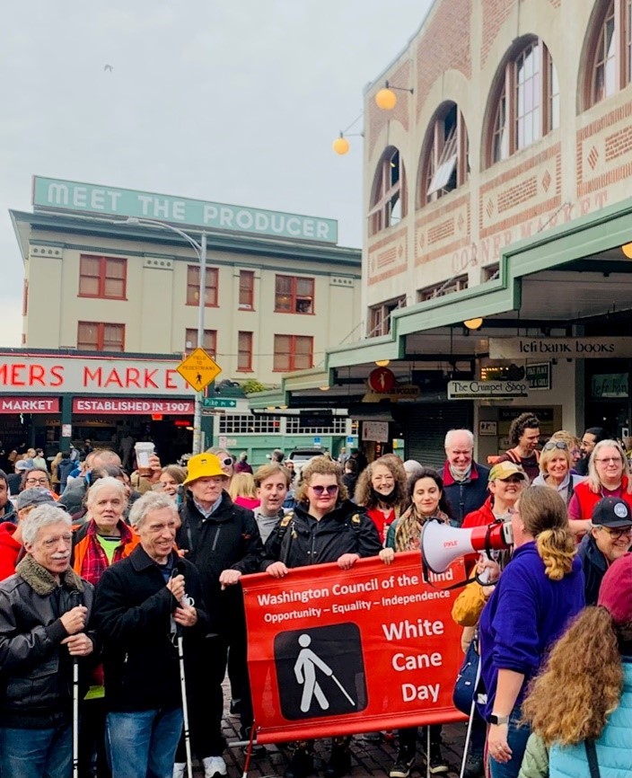 A group hold a banner announcing "White Cane Day" on behalf of the Washington Council of the Blind in front of the iconic Pike Place Market.